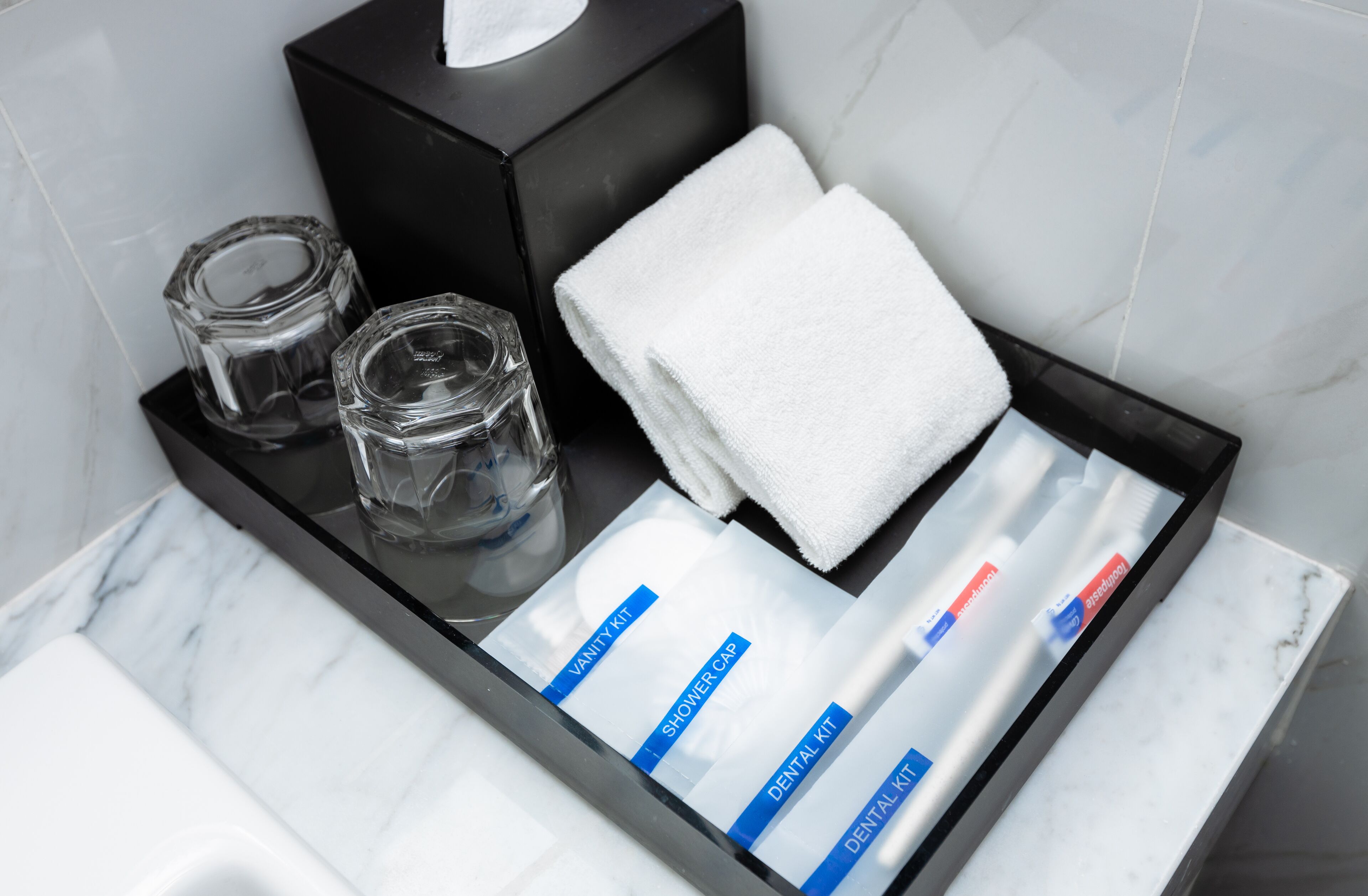 Un meuble-lavabo de salle de bain avec un plateau noir sur lequel sont posés une serviette blanche, deux récipients en verre et un ensemble de produits de toilette dans un étui transparent.