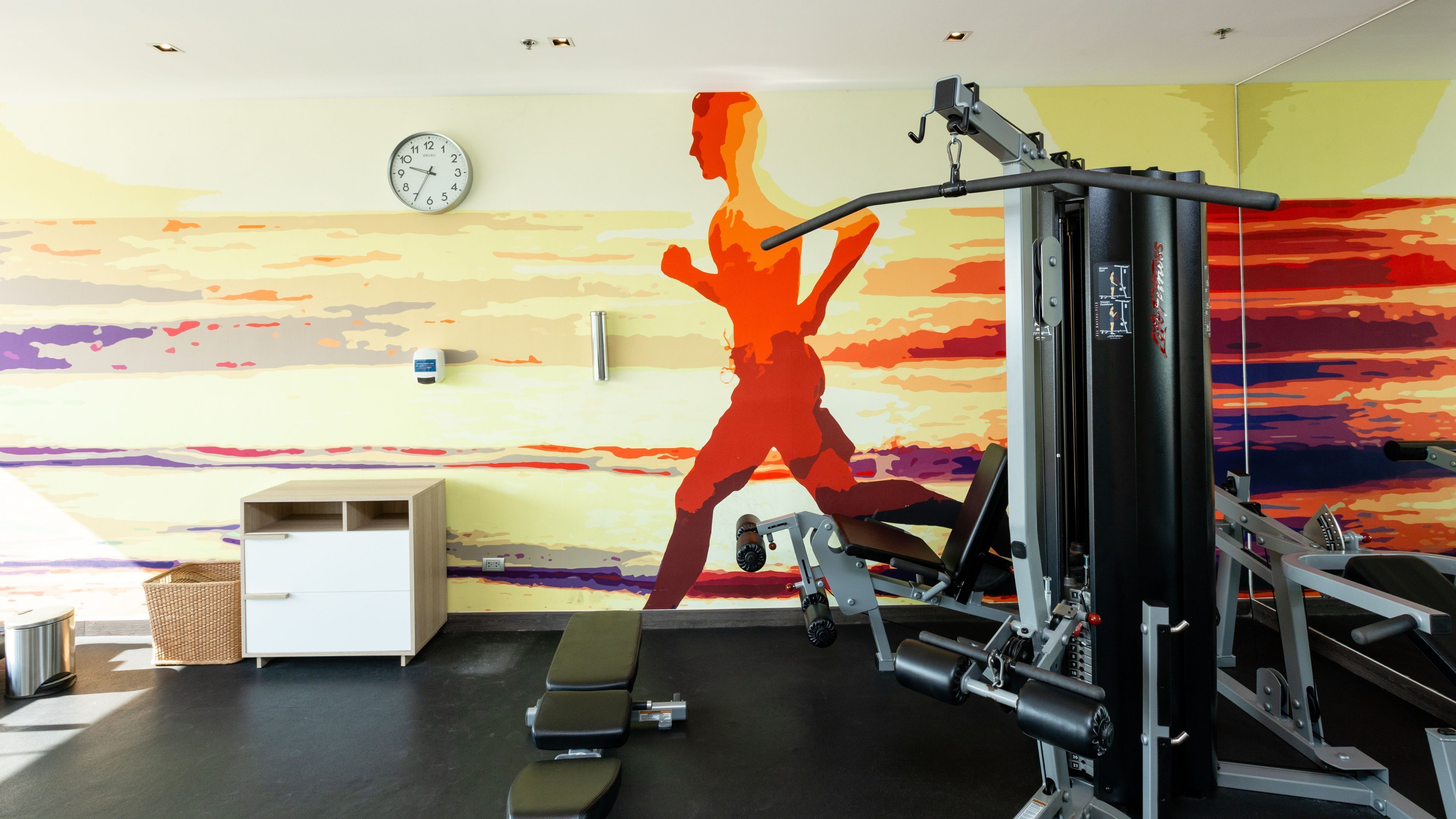 Ein Fitnessraum mit Trainingsgeräten, einer Wanduhr und einem großen Wandbild einer laufenden Person.