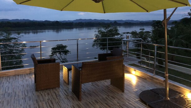 Una terraza con mobiliario de mimbre y una sombrilla amarilla, con vistas a un cuerpo de agua y montañas distantes.