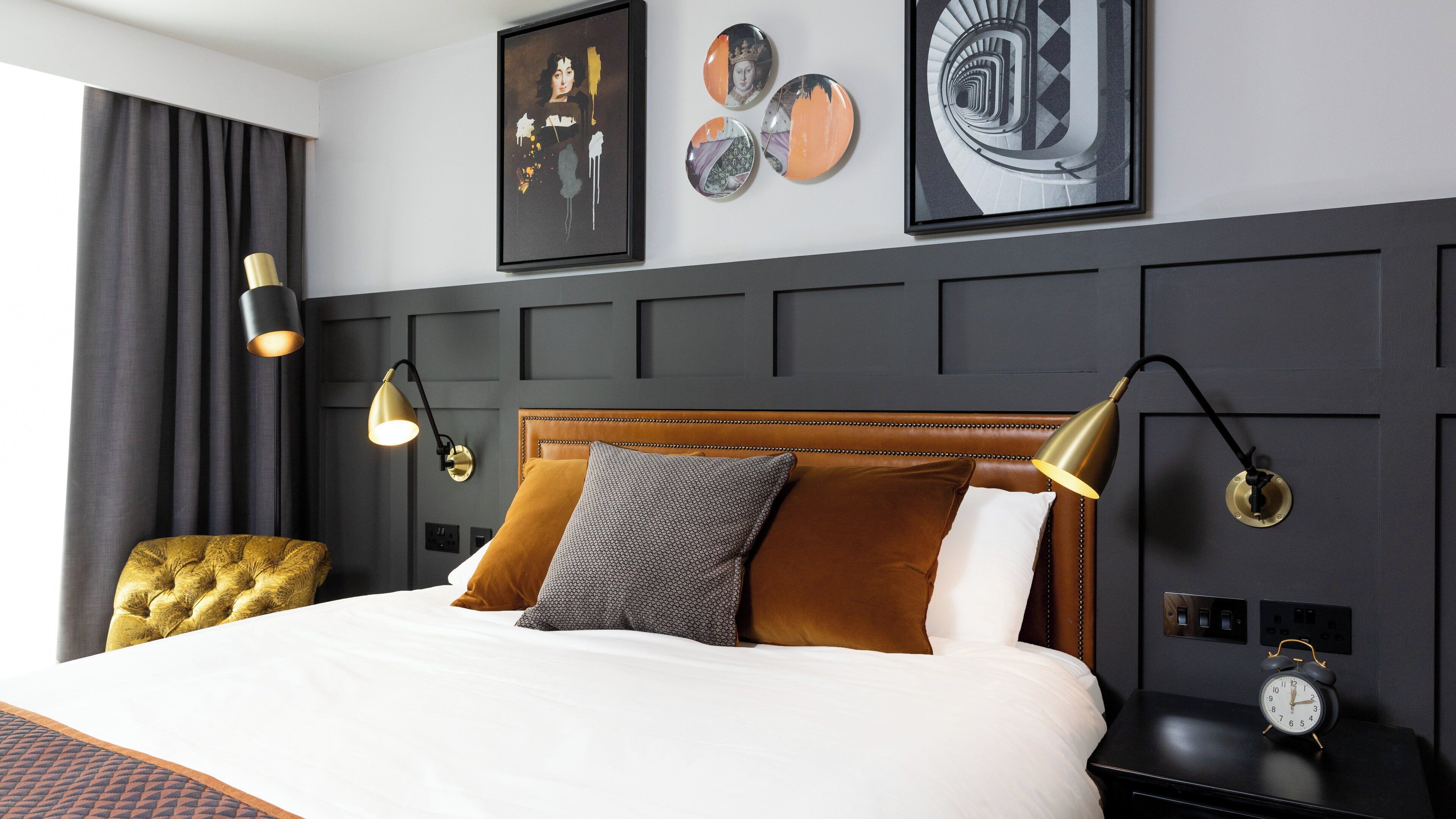 A hotel room with a large bed, two wall-mounted lamps, a clock, and artwork on the wall.