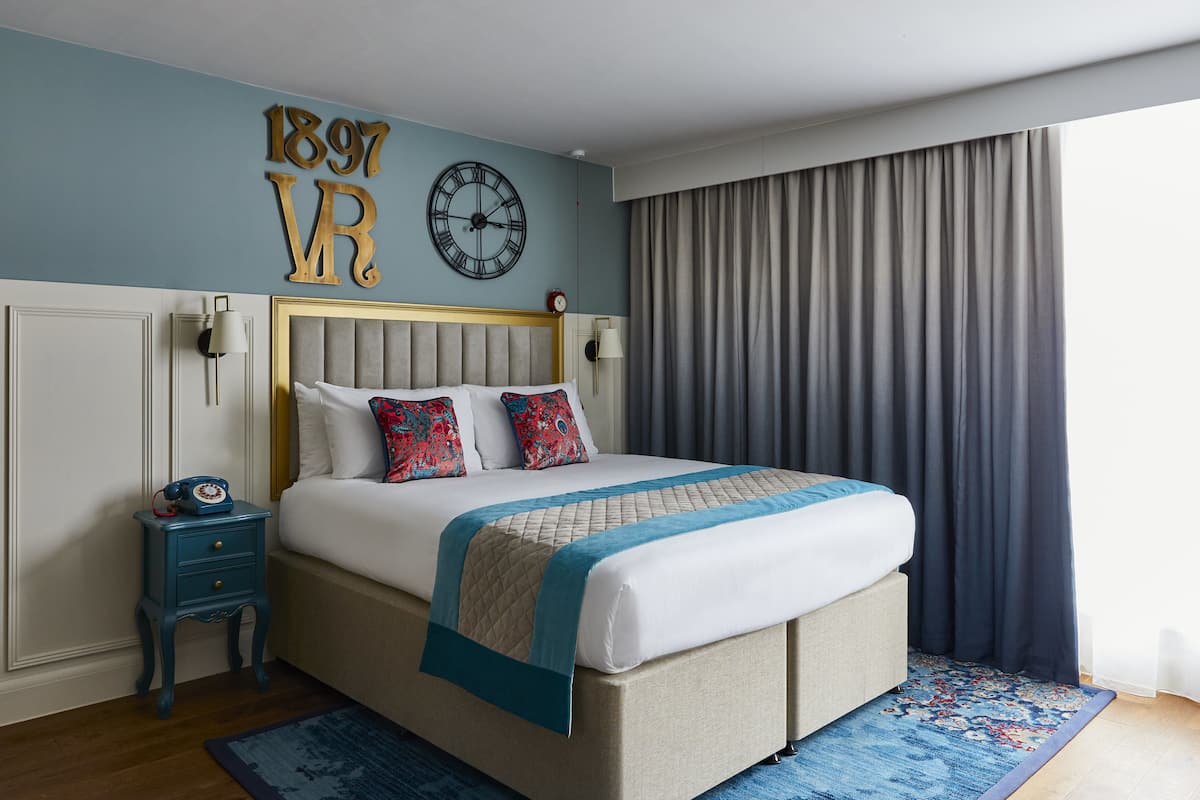A neatly made bed with a headboard, a bedside table with a lamp, and a wall clock.