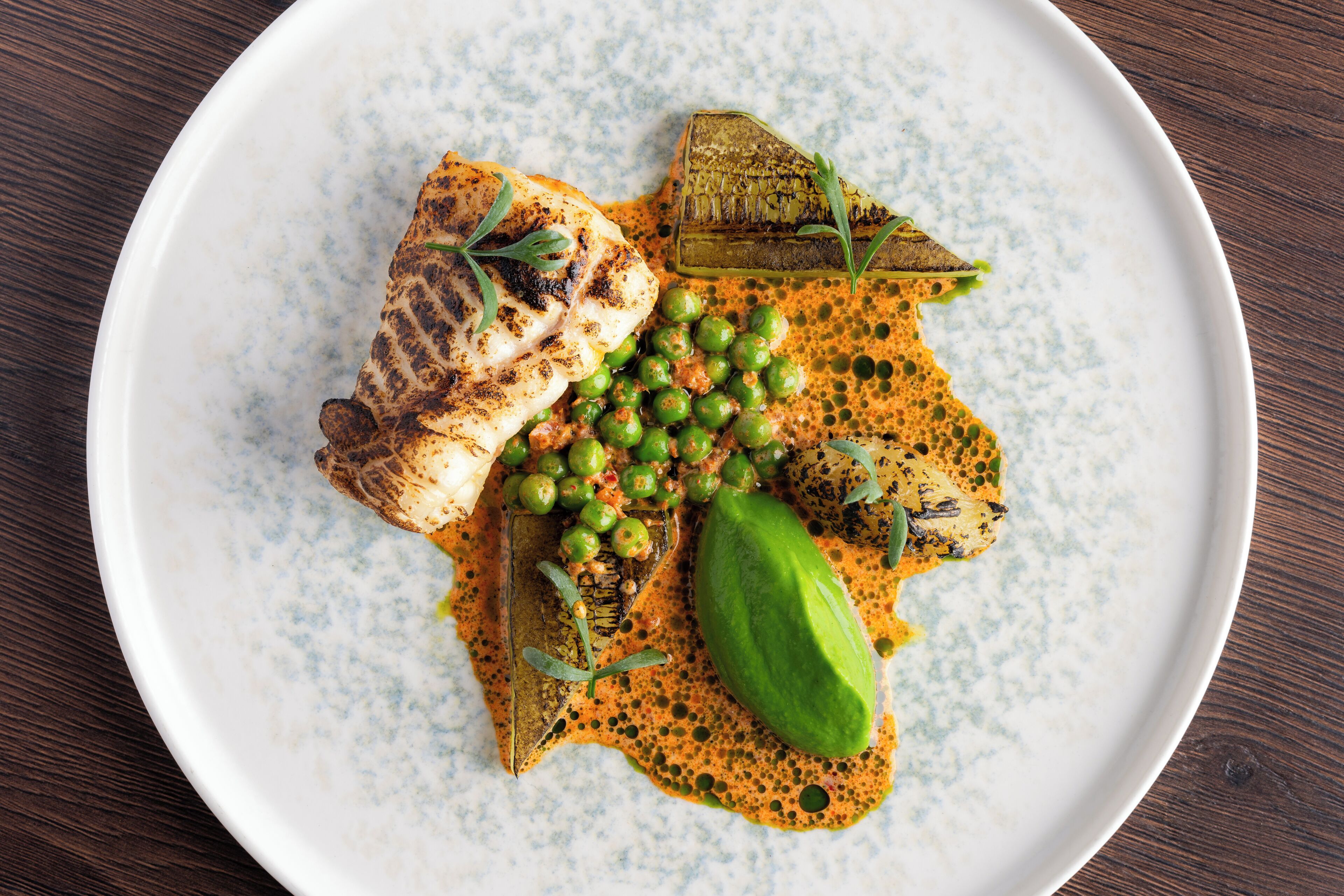 Un plato de comida con pescado a la parrilla, guisantes y una verdura verde sobre un plato moteado.