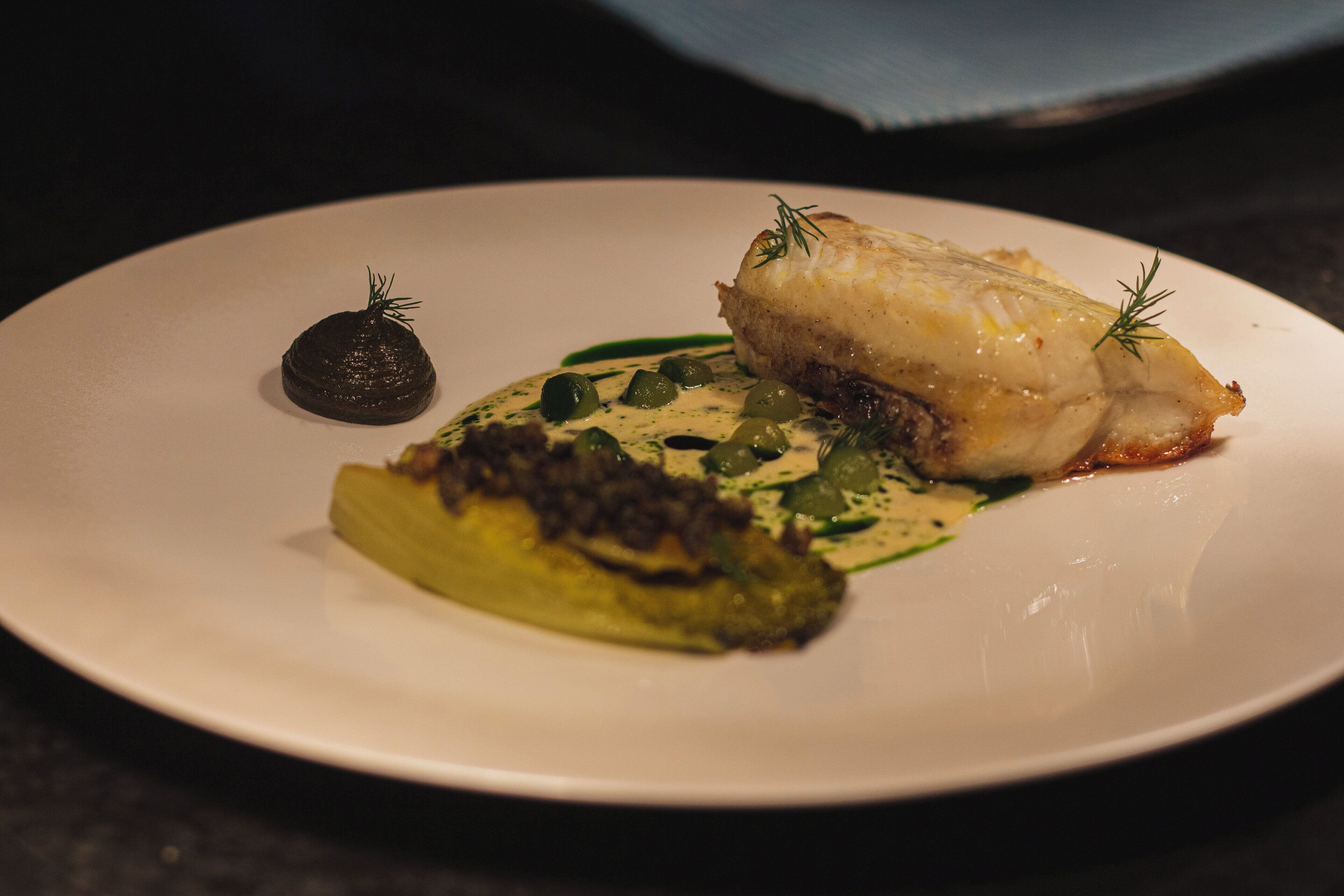Une assiette contenant un morceau de poisson, un objet sphérique de couleur sombre et des légumes verts.