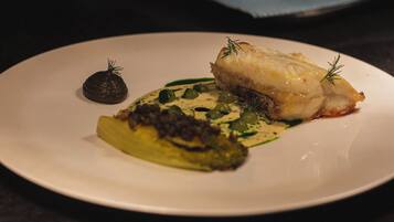 Un plato con un trozo de pescado, un objeto esférico oscuro y verduras verdes.