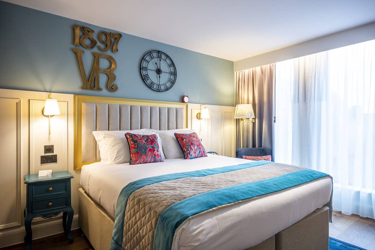 A hotel room with a large bed, two bedside tables with lamps, a wall clock, and a large window with curtains.