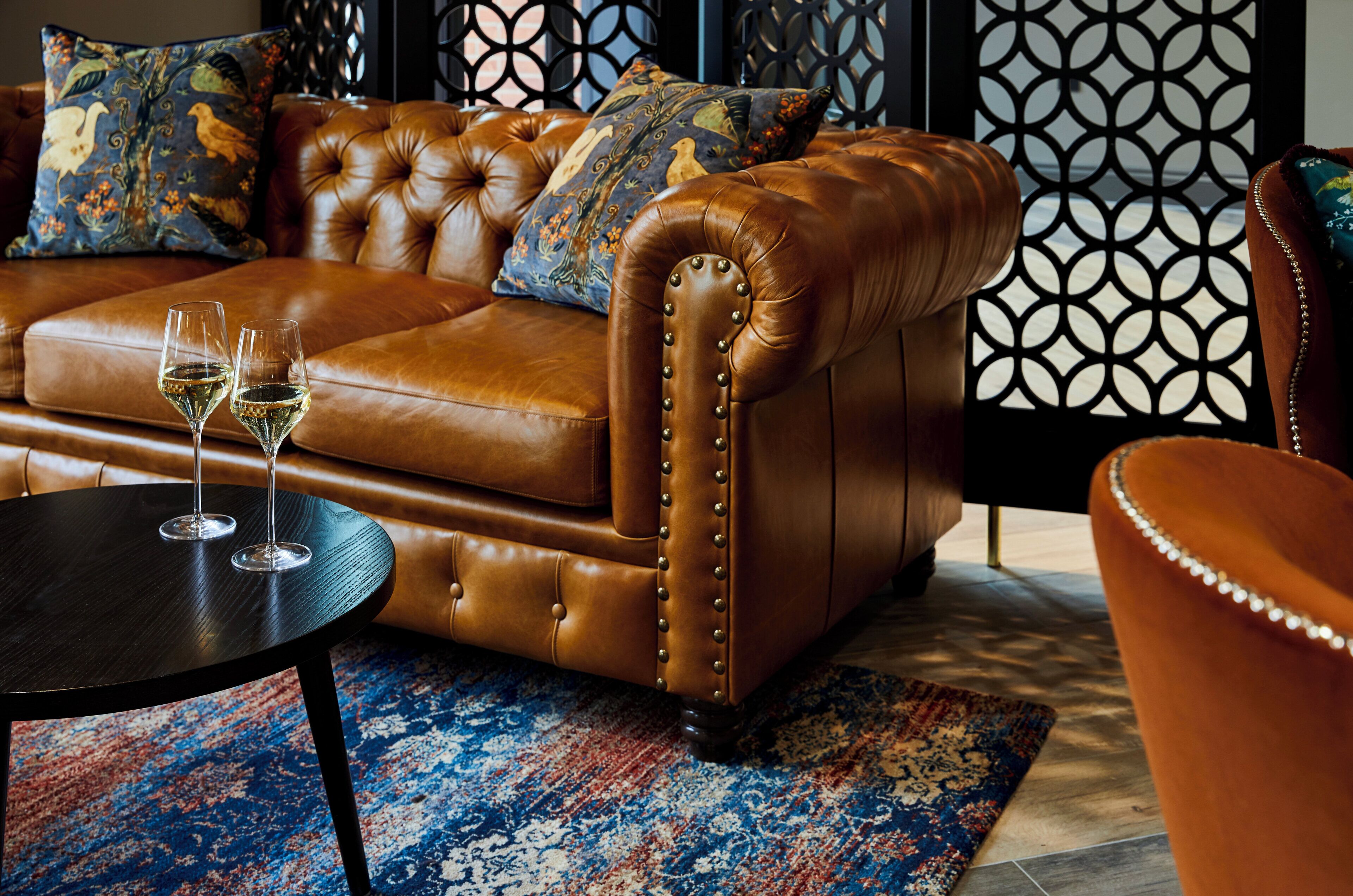 A brown leather sofa with tufted detailing and decorative buttons, accompanied by matching armchairs and a small round table with two wine glasses.