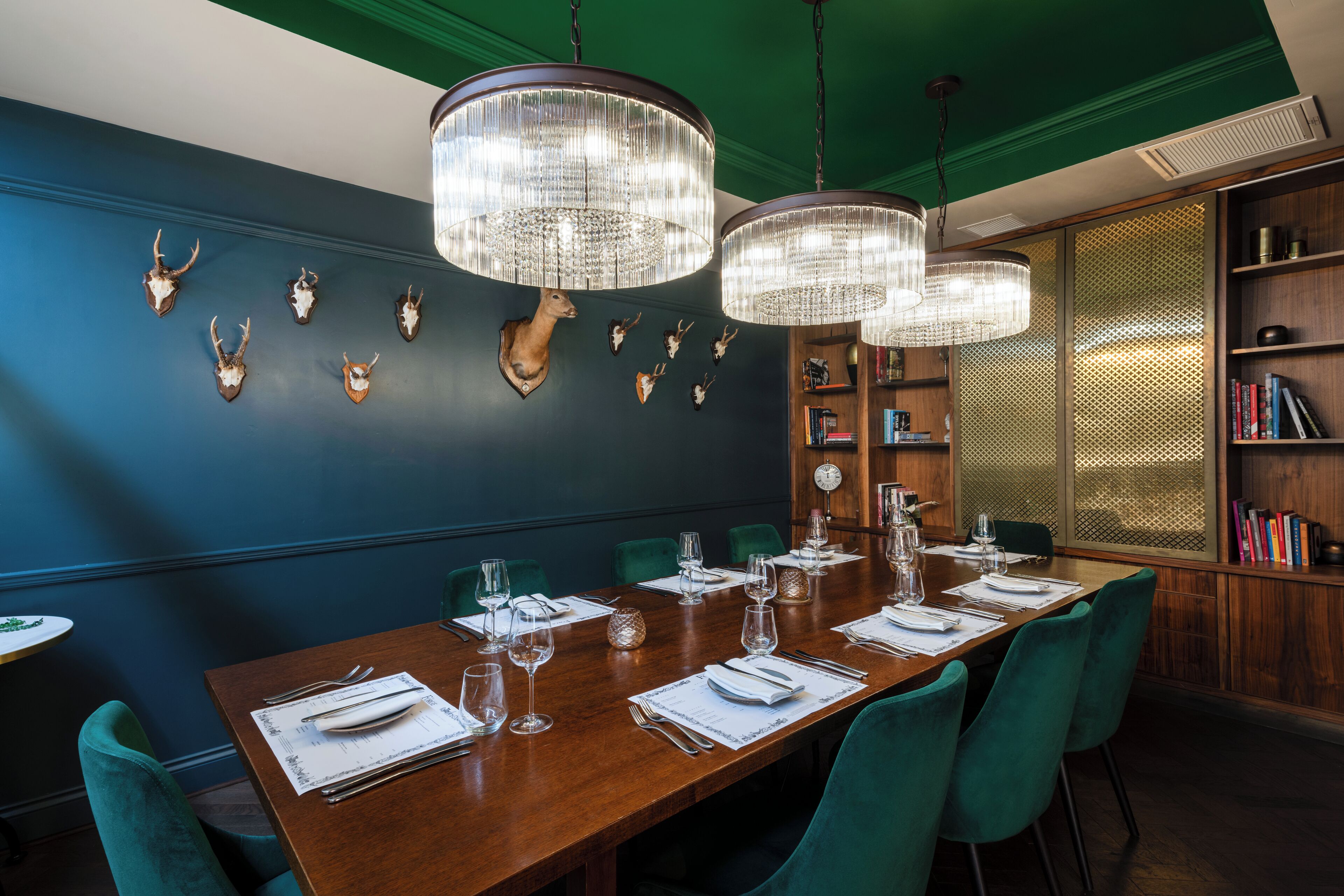 Un espace repas avec une longue table en bois, dressée pour six personnes, entourée de chaises turquoise et éclairé par trois grands lustres en cristal.