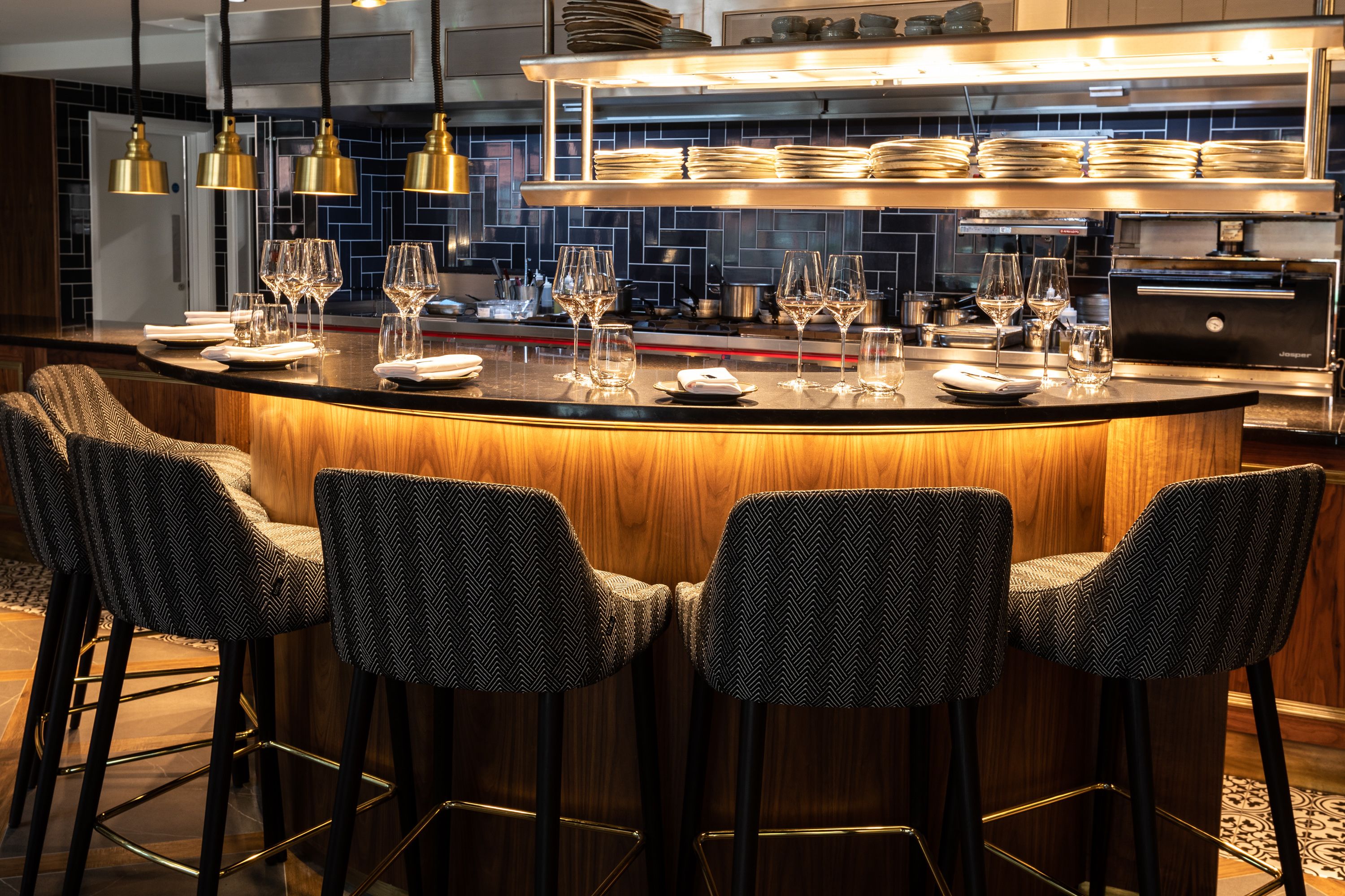 Uma área de bar com balcão de madeira, quatro banquetas estampadas, e uma cozinha ao fundo com pratos e copos empilhados.