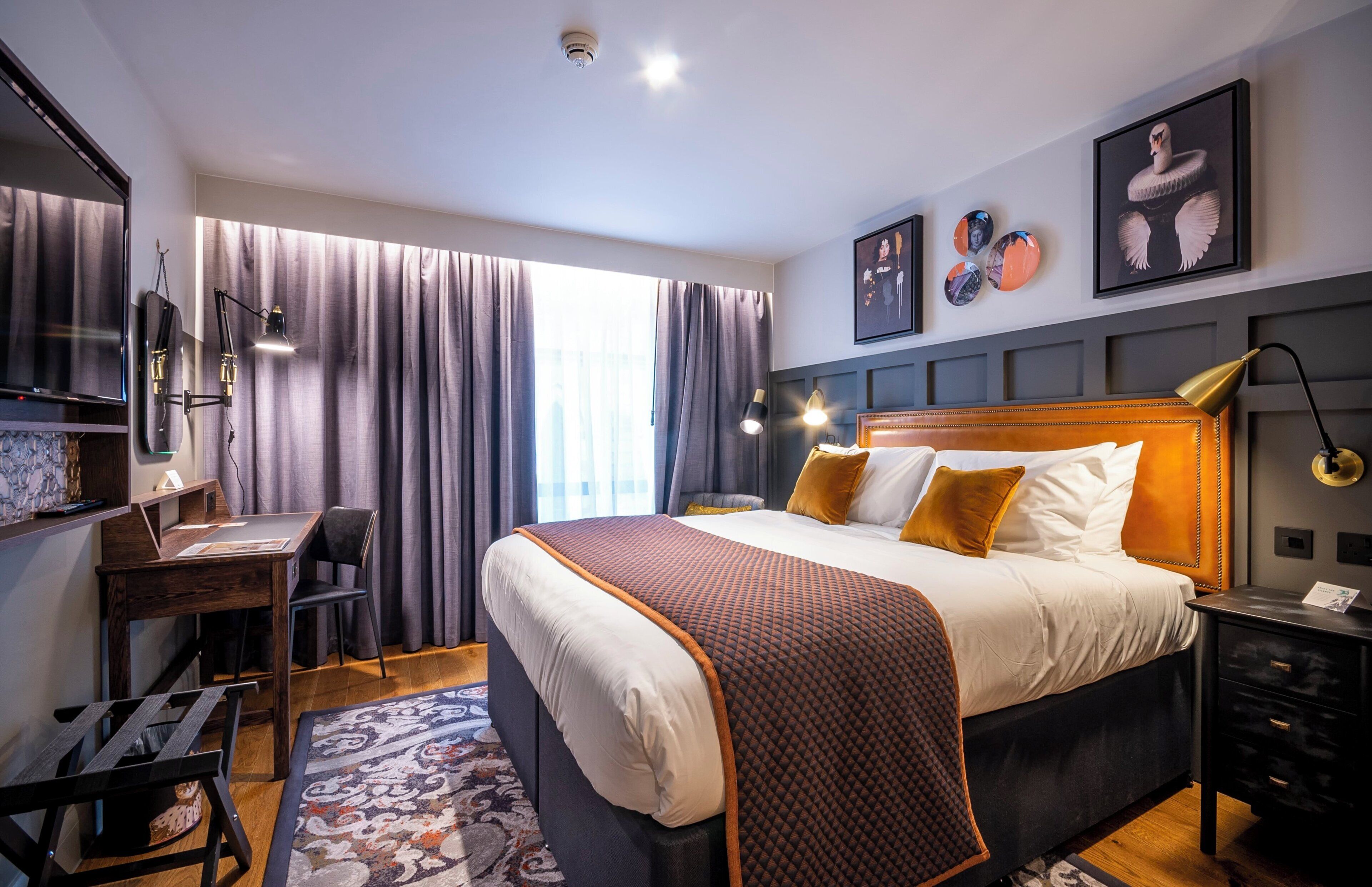 A modern hotel room with a large bed, a desk, a chair, and a desk lamp.