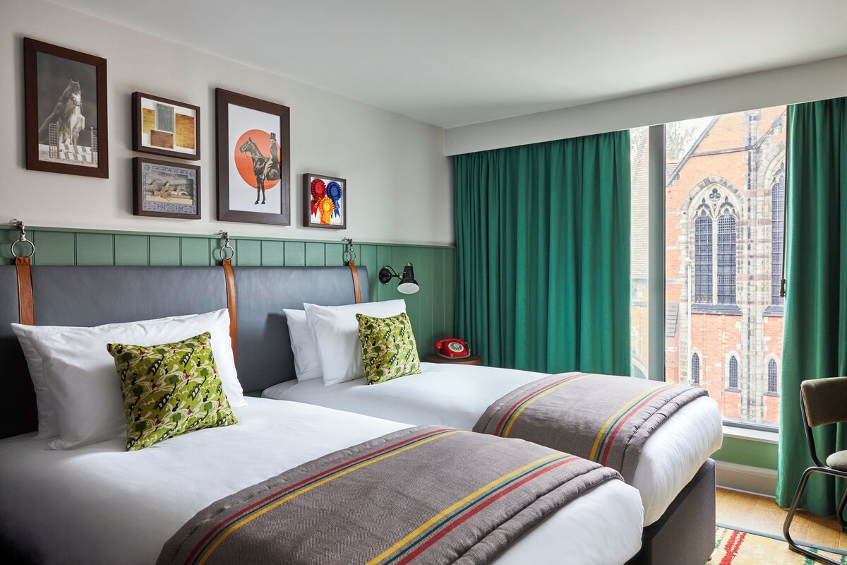 A hotel room with two beds, a green curtain, a red telephone, and a view of a brick building.