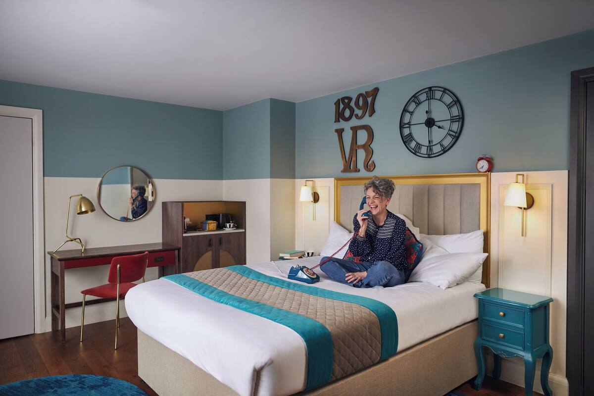 A hotel room with a bed, a desk, a chair, a nightstand, a clock, and a mirror.