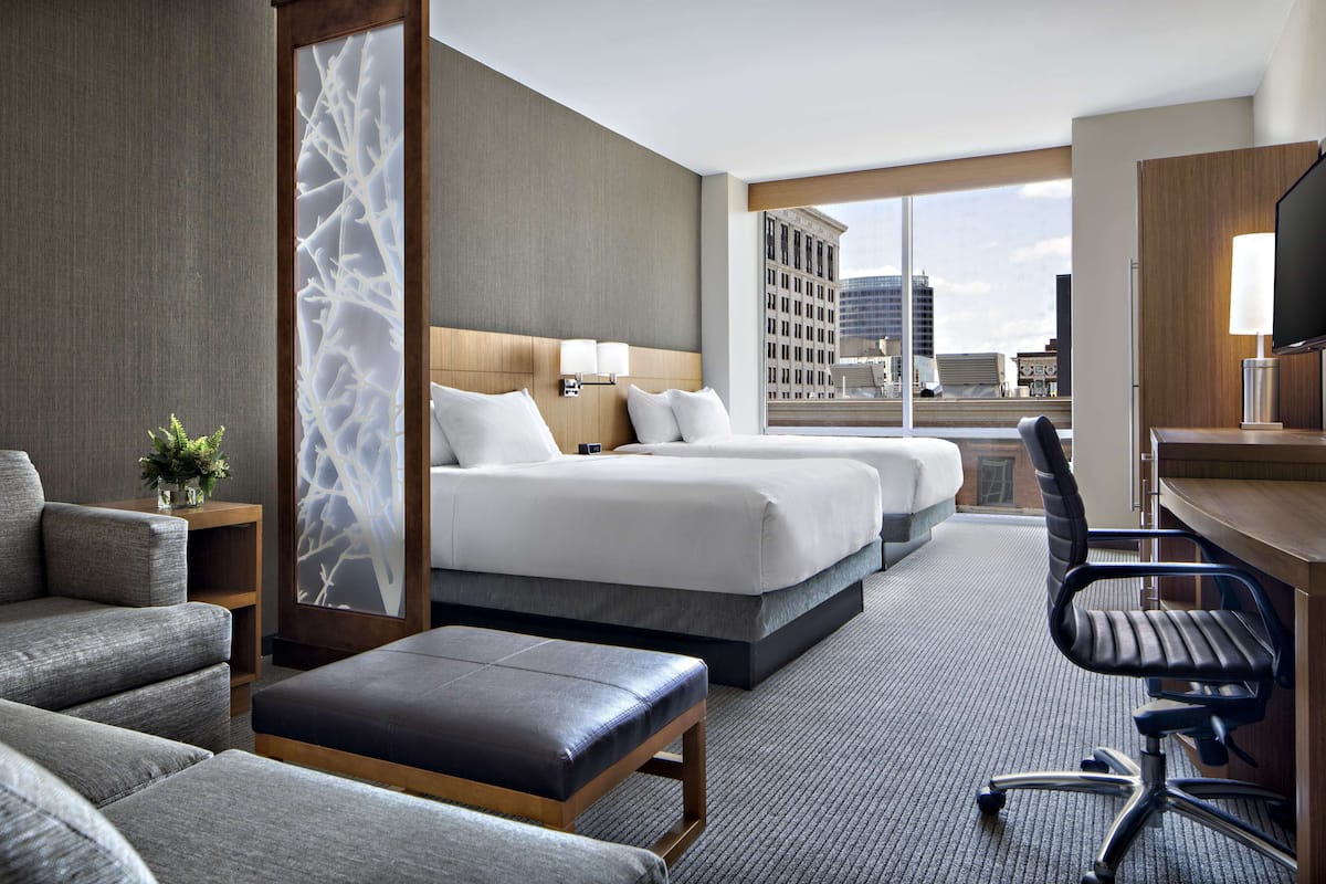 A hotel room with two beds, a desk, a chair, and a large window offering a city view.