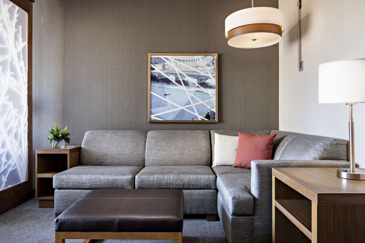 A modern living room with a grey sofa, a wooden side table with a plant, a framed picture on the wall, and a lamp on a wooden side table.