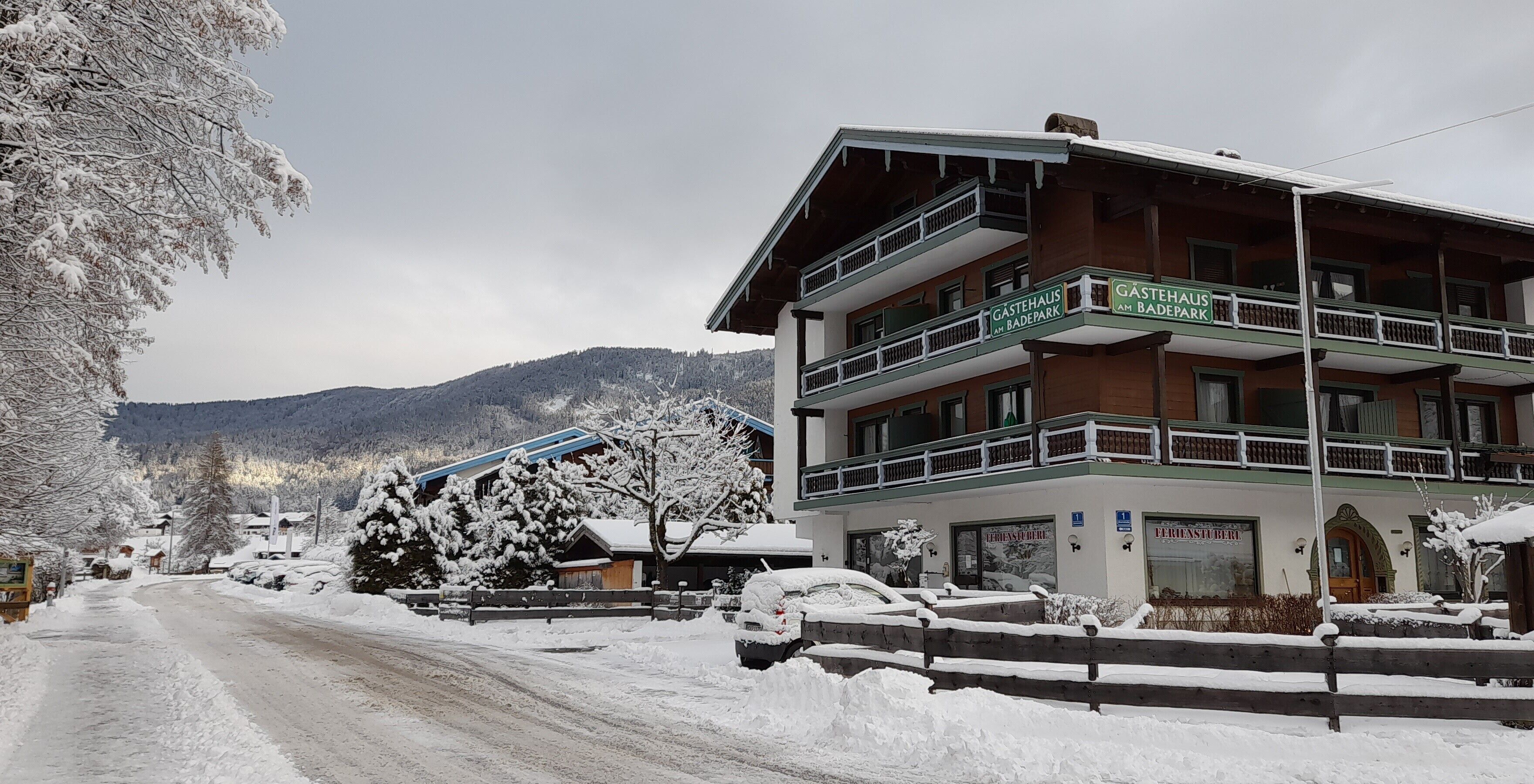 A multi-story building with balconies, a sign reading "Gastehaus am Badepark," and a snow-covered landscape in the background.