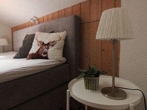A bedroom with a bed, a grey headboard, a lamp, a plant, and a pillow with a deer design.