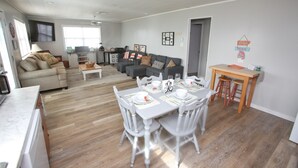 A spacious living area with a dining table set for four, a grey sofa, and a kitchen area with a TV. - Charming 2nd Row remodel with Beach Views (Surfside Beach)