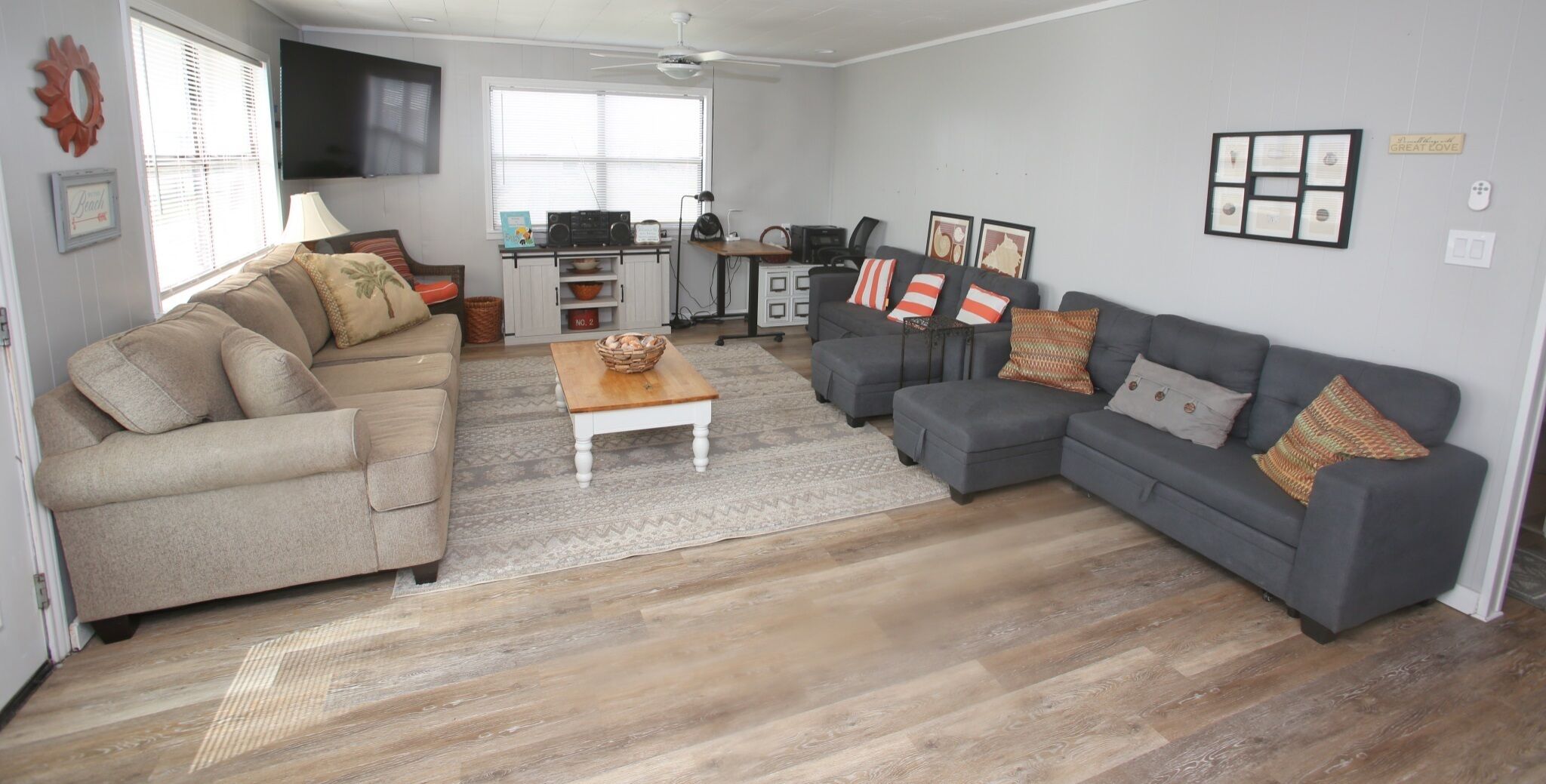 Amplia sala de estar con un sofá gris, un sofá color beige y una mesa de centro de madera. En el fondo, se observa una zona de cocina con un televisor montado en la pared.
