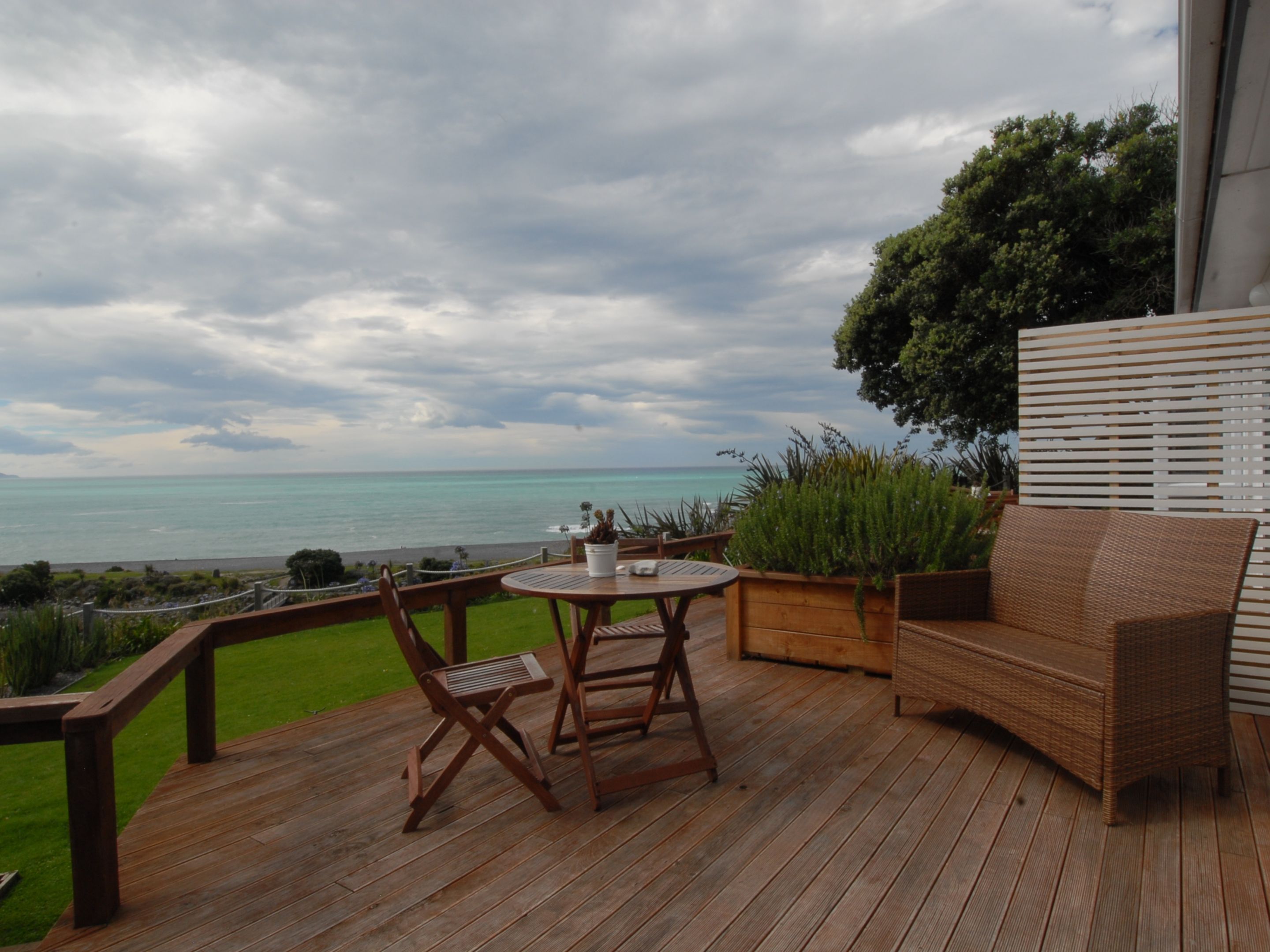 A wooden deck with a round table and two chairs, overlooking a beach and the ocean.