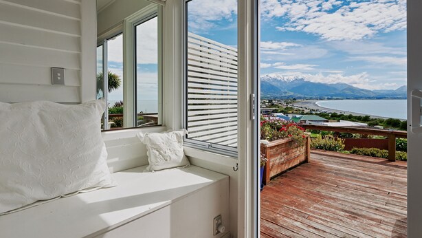 A room with a large window, a white cushioned bench, and a view of a lake and mountains.
