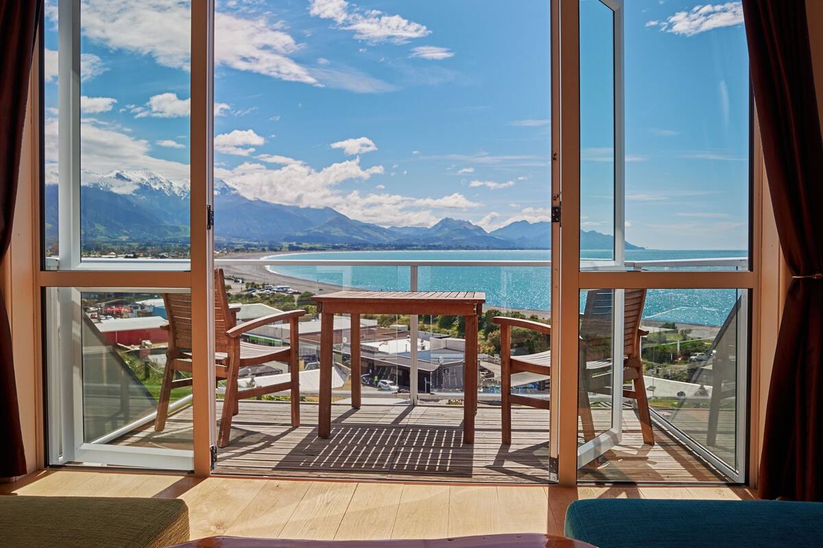 A balcony with a wooden table and chairs, overlooking a coastal area with mountains and a clear sky.