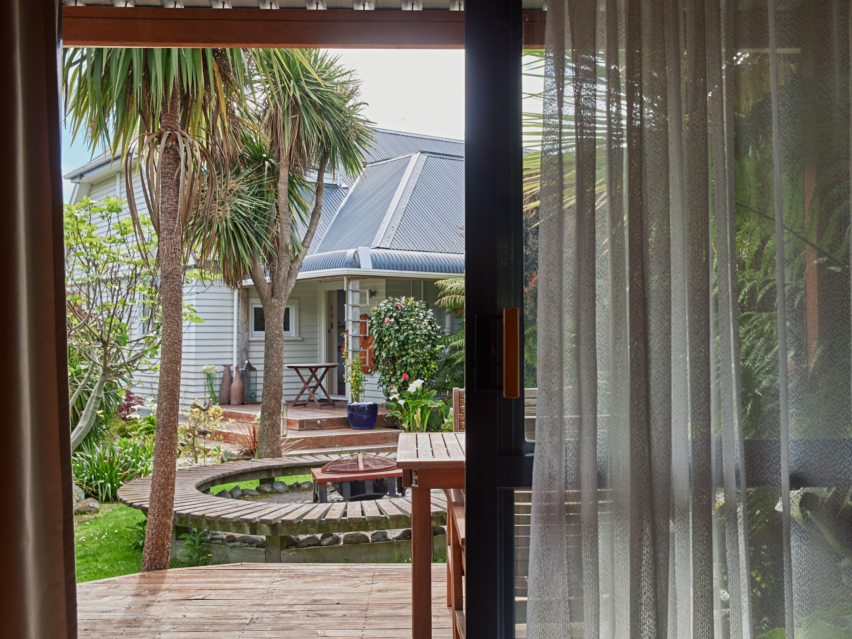 Una terraza de madera con una mesa y sillas, rodeada por un jardín con una palmera y una casa al fondo.