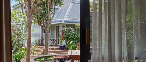 A wooden deck with a table and chairs, surrounded by a garden with a palm tree and a house in the background.