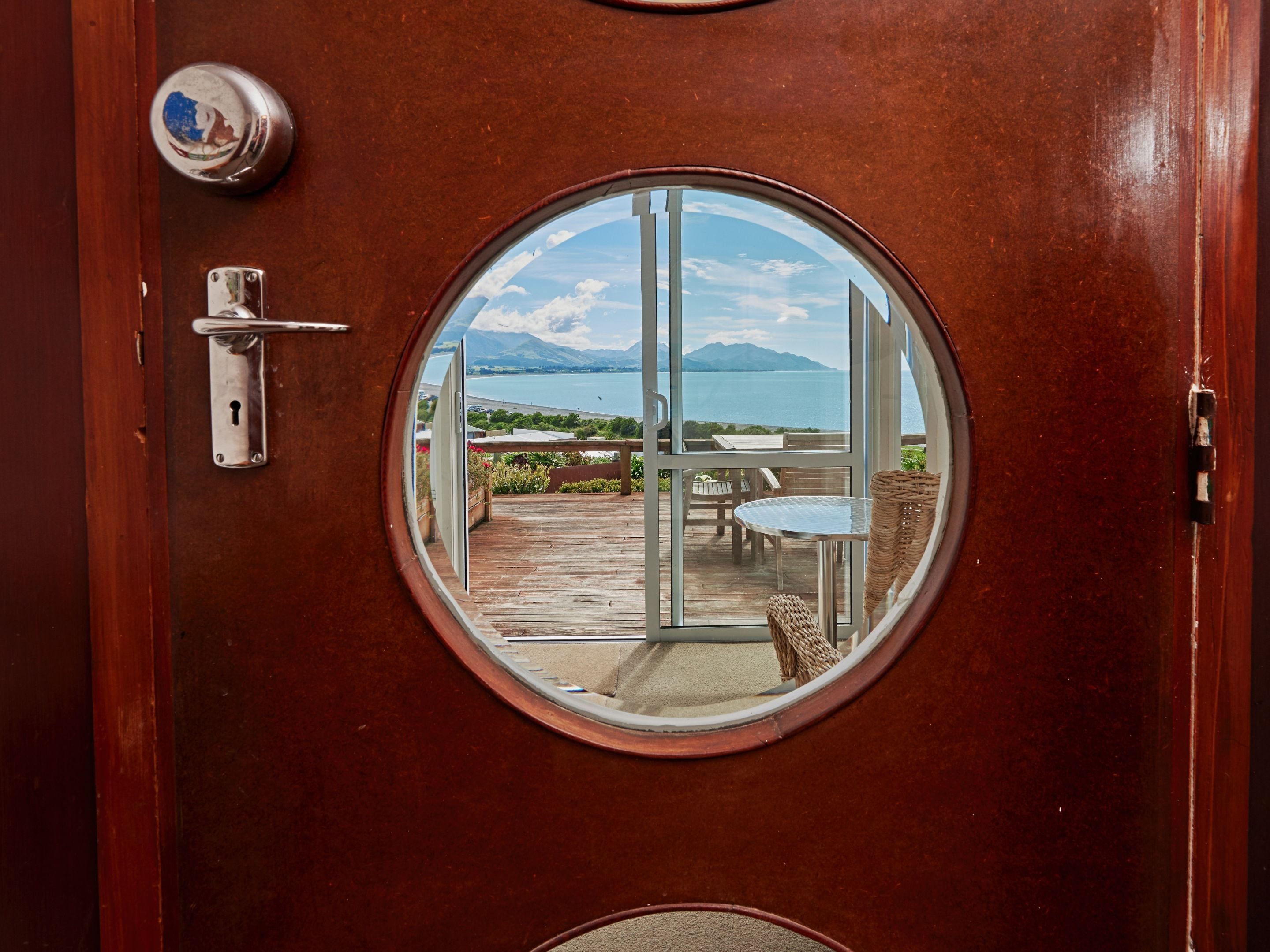 Una puerta de madera con un ventanal circular que muestra una terraza con mobiliario exterior y vistas a un cuerpo de agua y montañas.