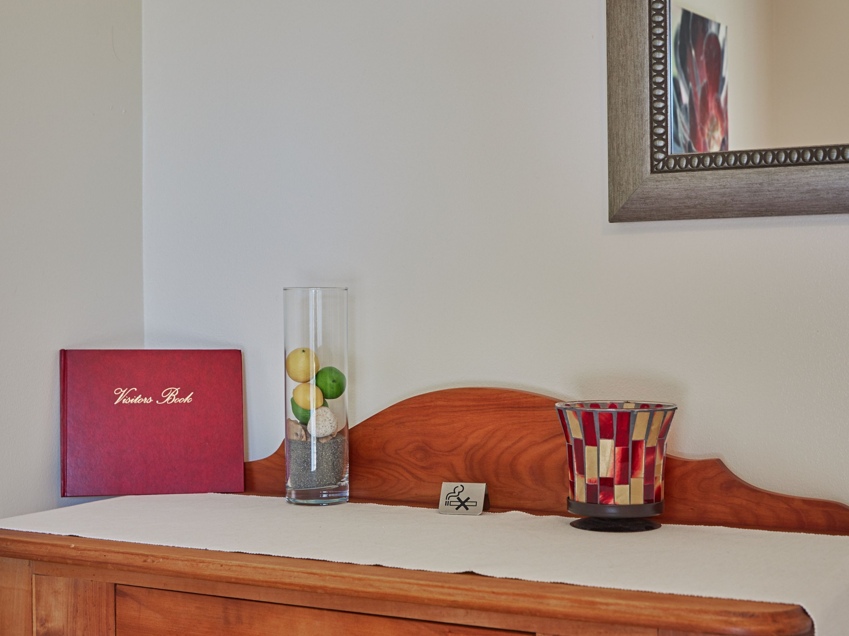 A wooden headboard with a framed mirror, a red book titled "Visitors Book," a clear vase with fruits and a candle, and a striped candle holder.