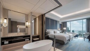 Habitación de hotel moderna con una bañera ovalada grande, una cama doble, zona de estar con una silla y un ventanal con vistas.