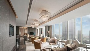 Un moderno vestíbulo de hotel con techo alto, amplias ventanas que ofrecen vistas de la ciudad, un área central de asientos con sofás y sillones, y un televisor de pantalla plana montado en la pared.