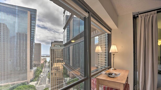 A high-rise hotel room with a large window offering a city view, a small wooden table with a lamp, and a red-striped ottoman. - Downtown Honolulu Suite Near Ala Moana Beach! (Honolulu)