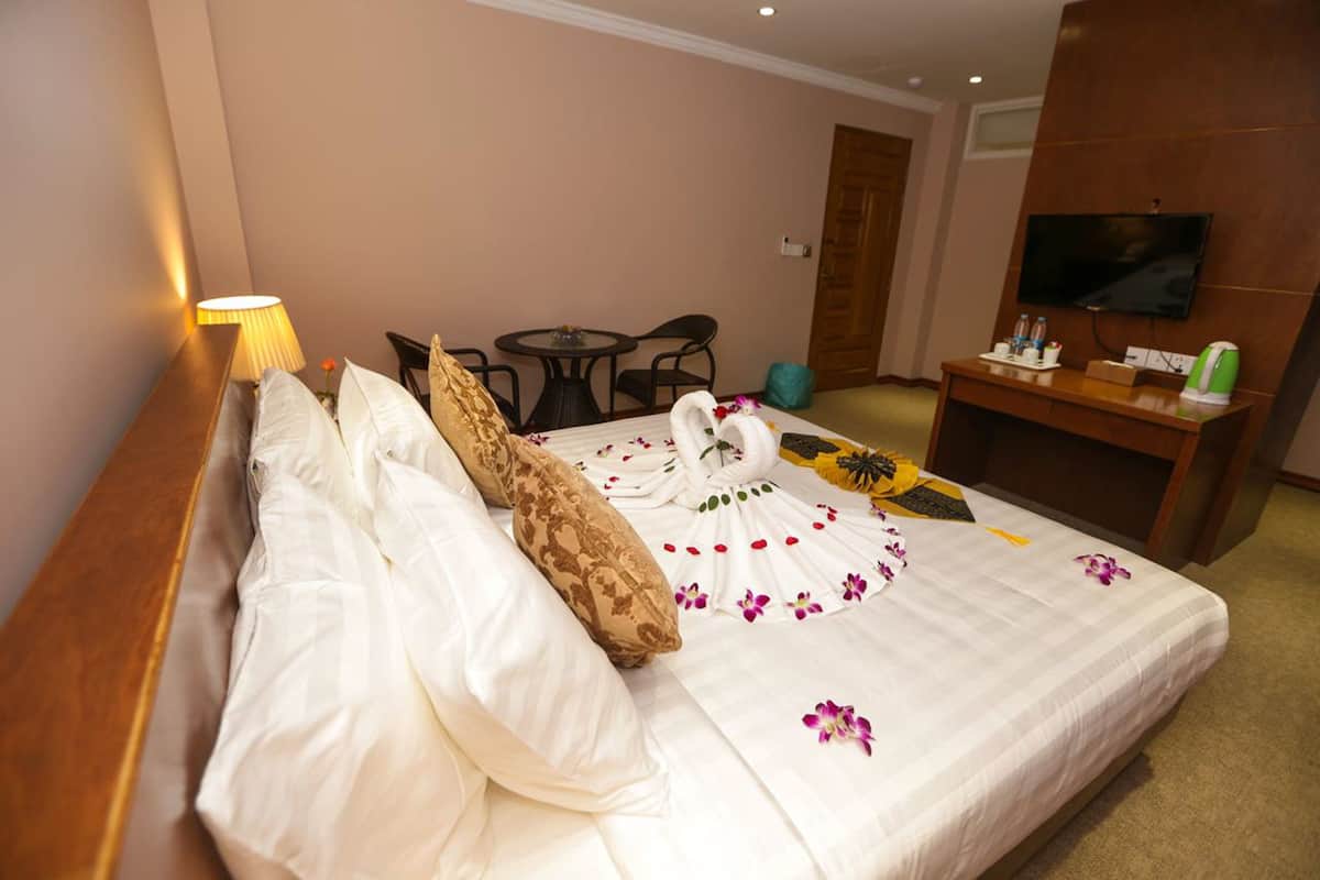 A hotel room with a neatly made bed, a small table with a chair, a television, and a desk with a kettle.