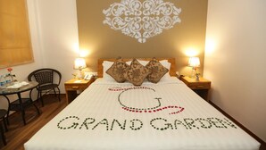 A hotel room with a bed featuring a decorative design, two bedside lamps, a small table with a chair, and a wall with a decorative pattern. - Grand Garden Hotel Yangon (Yangon)