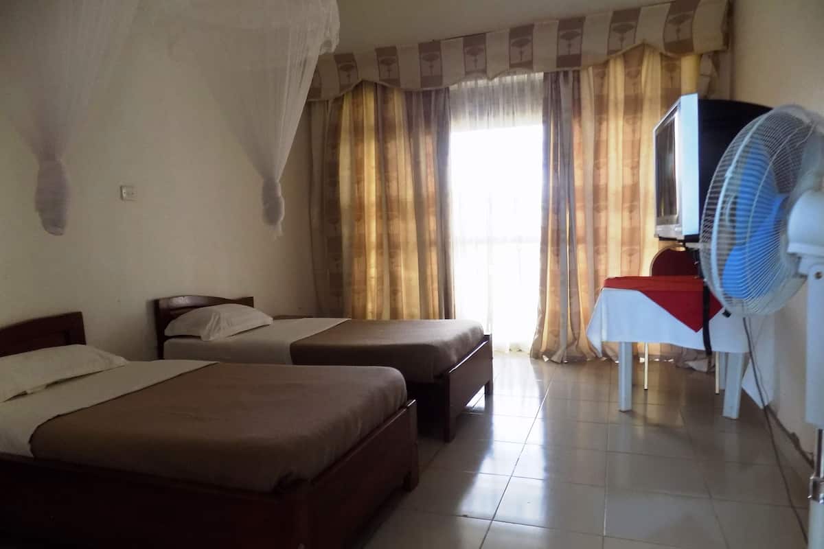 A hotel room with two beds, a desk with a chair, a standing fan, and curtains.