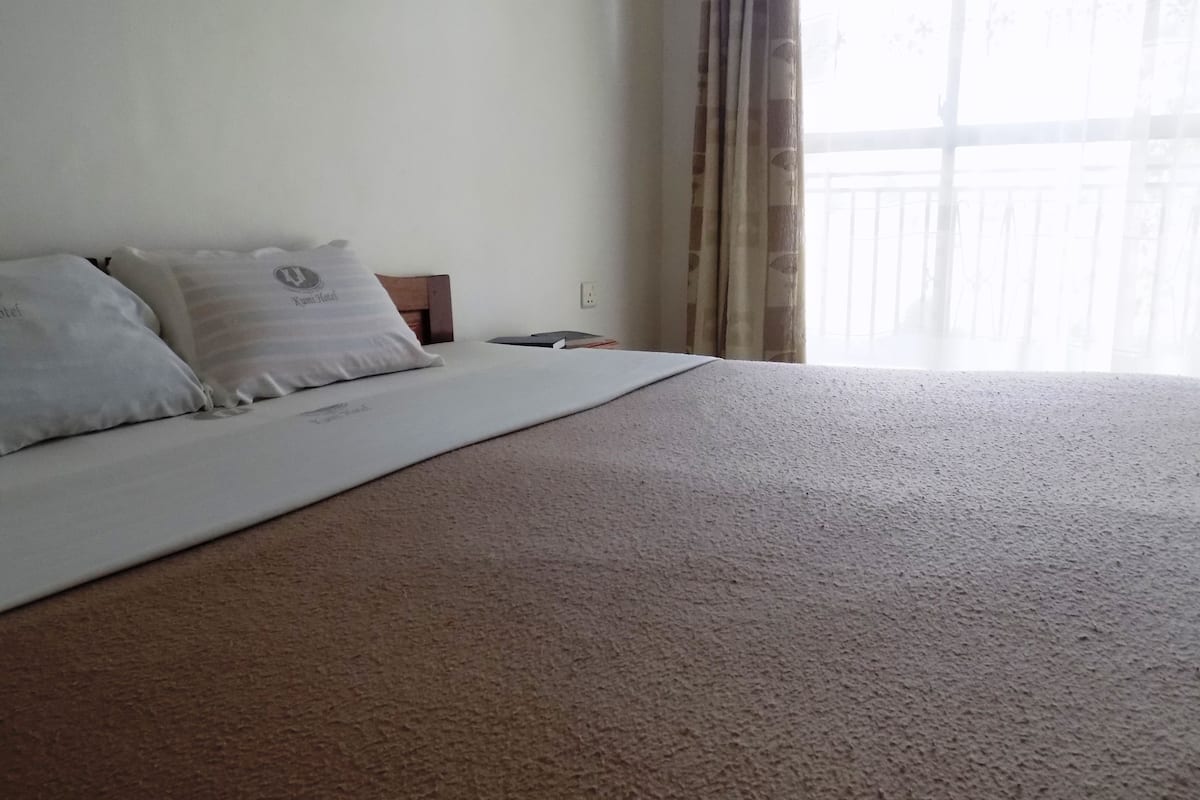 A neatly made bed with a large beige comforter and two pillows, a wooden nightstand with a book and a phone, and a window with curtains.