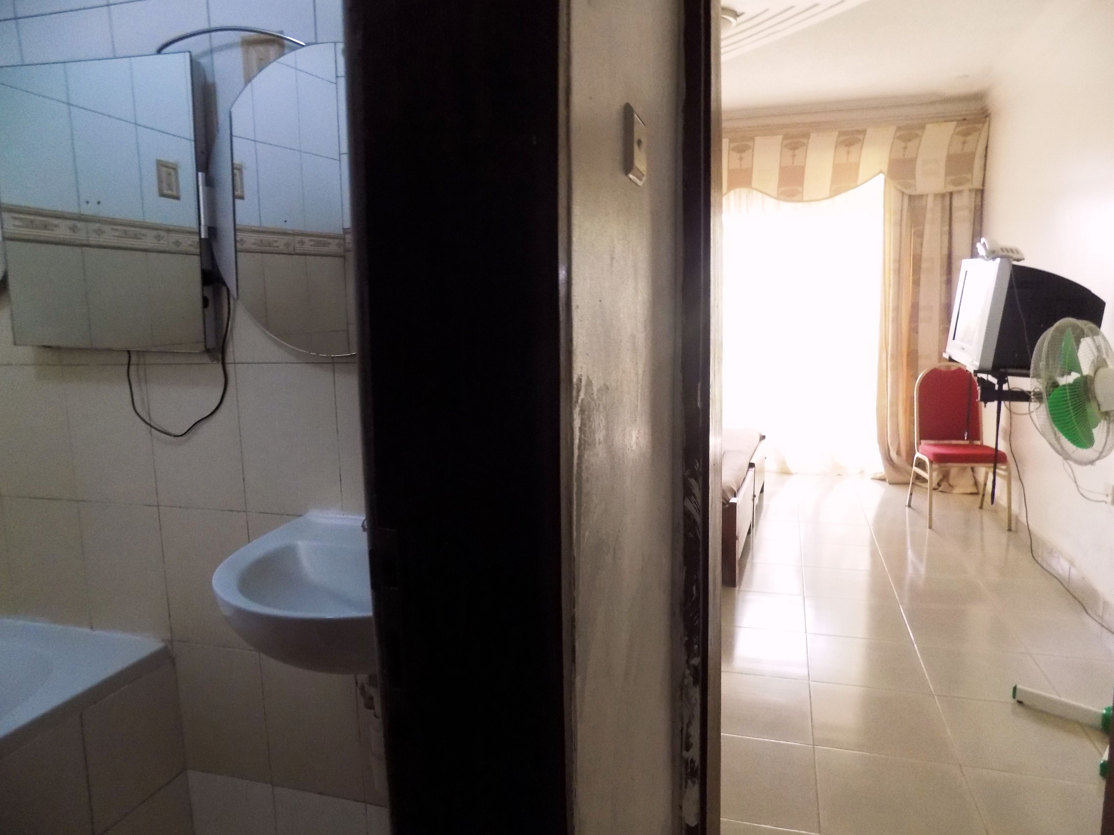 A bathroom with a sink, a mirror, and a toilet. There is a door leading to a room with a chair and a fan.