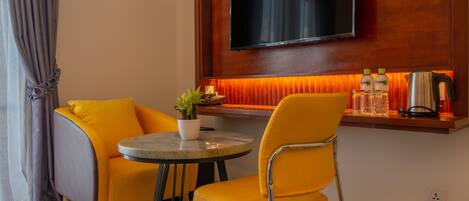 A room with a TV mounted on a wooden wall, a small table with a potted plant, two yellow chairs, and a coffee maker on a shelf.