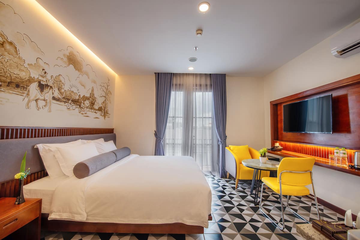 Une chambre d’hôtel avec un lit, une télévision, un coin repas avec des chaises jaunes et une grande fresque murale.