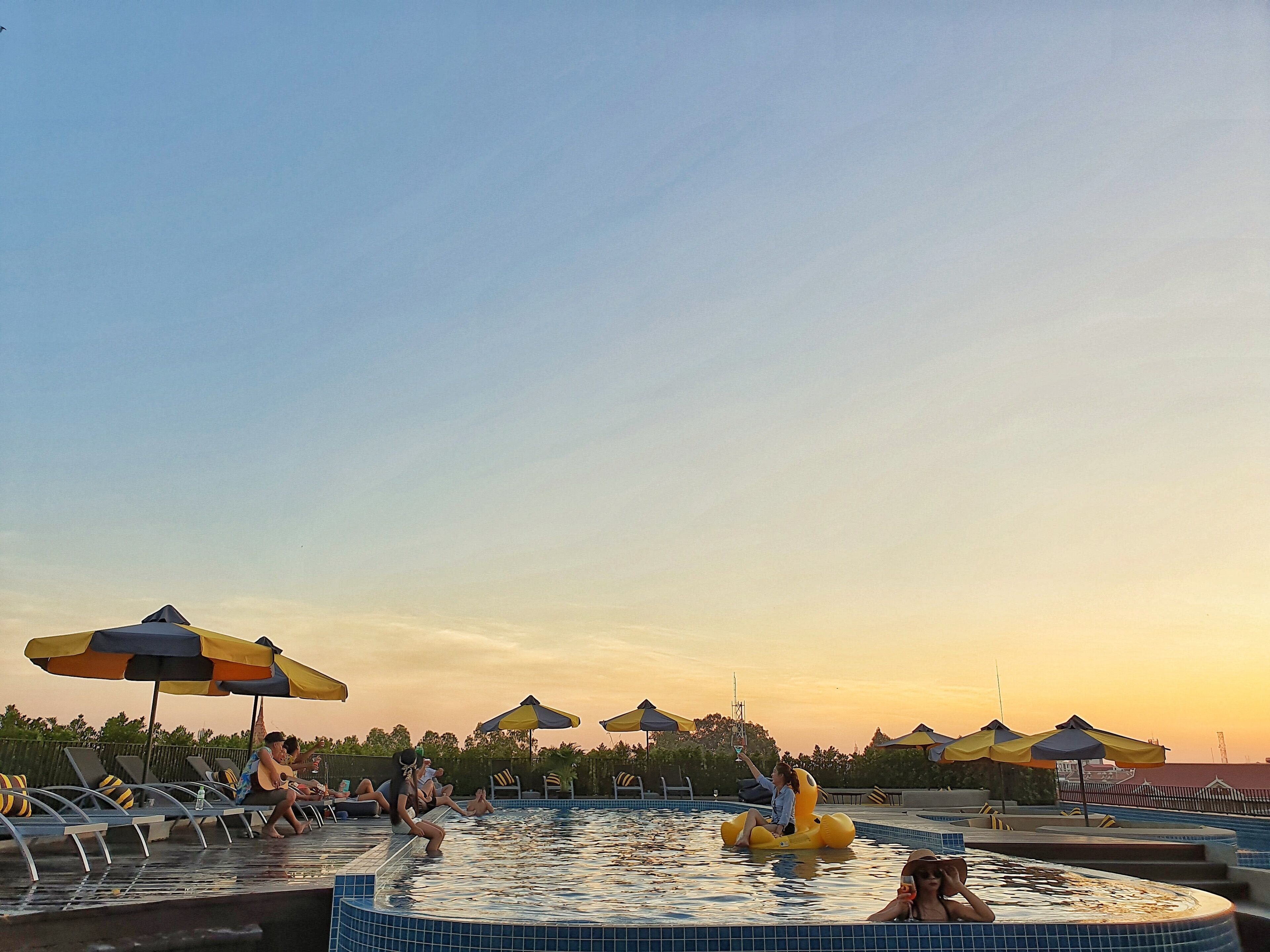 A pool area with lounge chairs and umbrellas, a hot tub, and a sunset in the background.