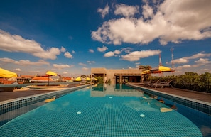 Una piscina con piso de azulejos azules, rodeada de sombrillas y sillas de descanso, bajo un cielo parcialmente nublado.
