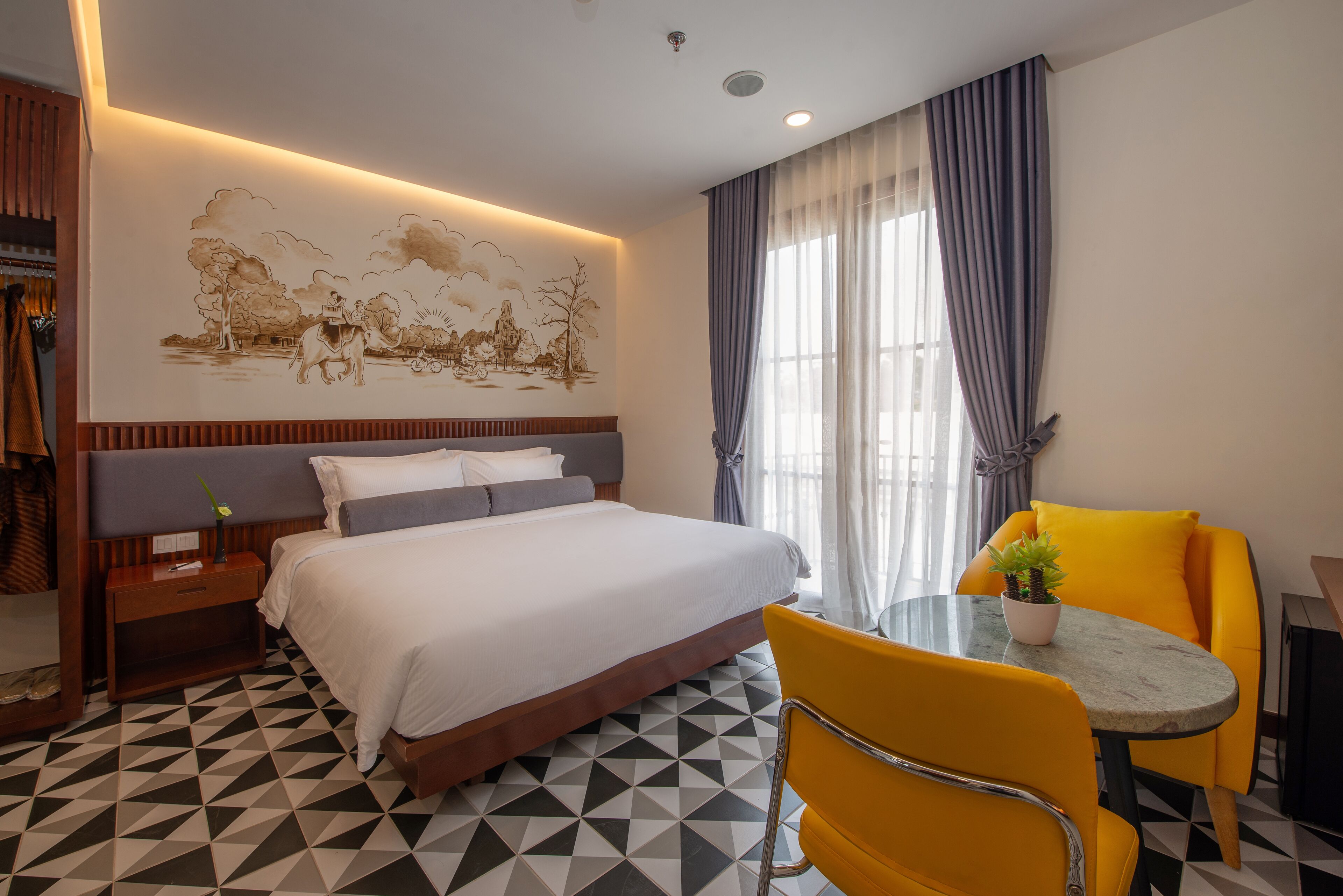 A hotel room with a bed, a bedside table, a chair, a small table with a plant, and a wall mural.