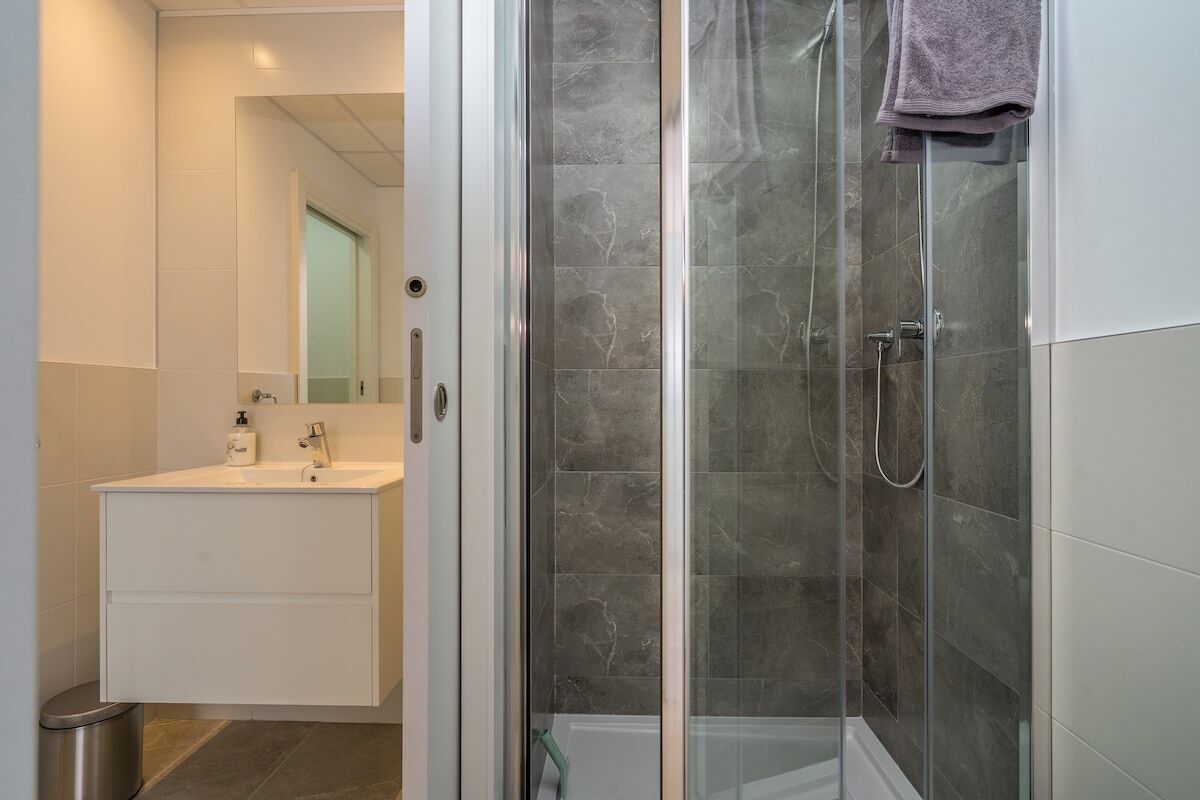 A modern bathroom with a glass-enclosed shower, a white vanity with a mirror, and a trash bin.