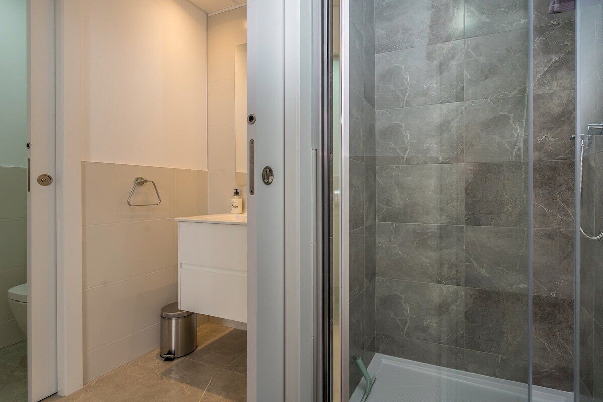 Een moderne badkamer met een douchecabine van glas, een witte wastafel en een kleine prullenbak.