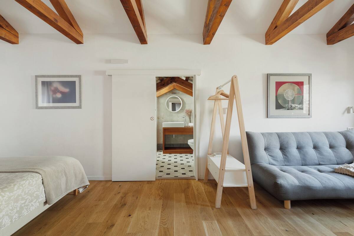 A bedroom with a bed, a grey sofa, wooden beams on the ceiling, and a bathroom visible through an open door.