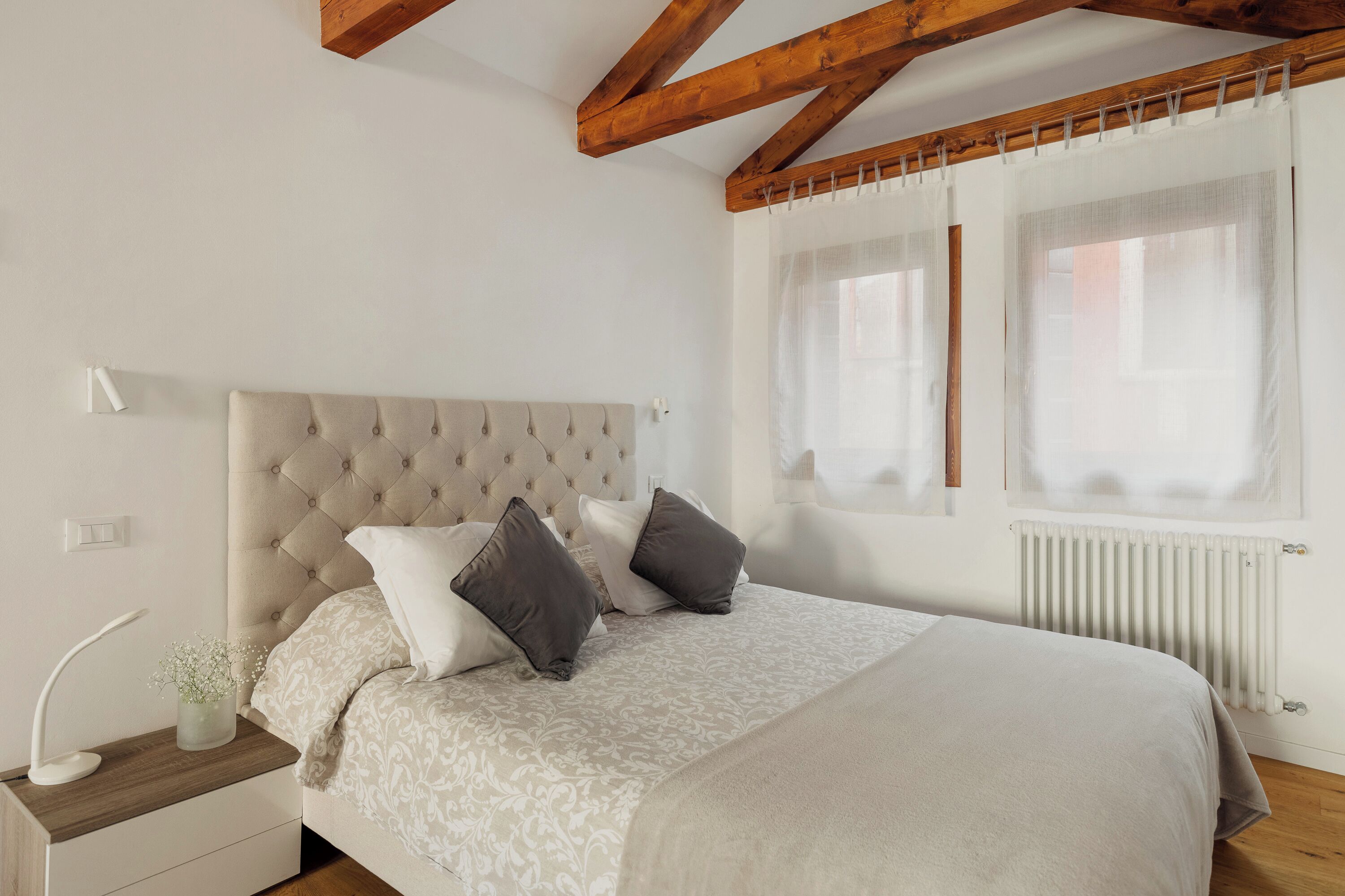 Un dormitorio con cabecera capitonada, una mesita de noche con lámpara y una ventana con cortinas traslúcidas.