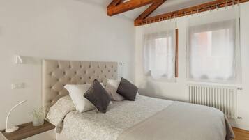 Un dormitorio con cabecera capitonada, una mesita de noche con lámpara y una ventana con cortinas traslúcidas.