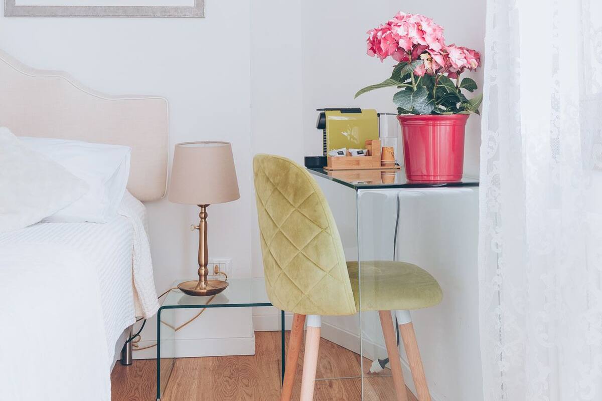 A bedroom with a bed, bedside table, lamp, chair, desk with a potted plant, and a framed picture on the wall.