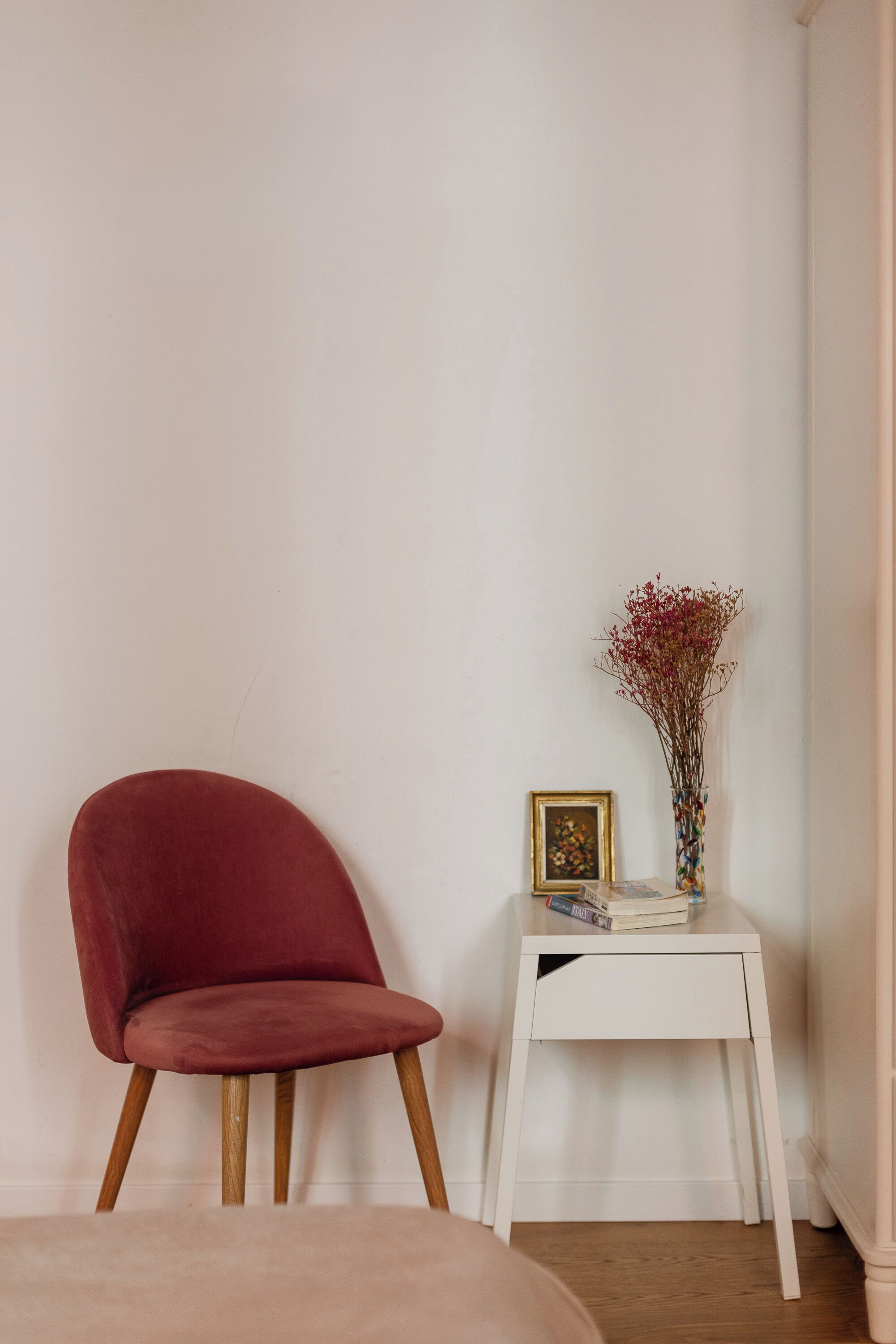 A red chair, a small white table with a vase and a framed picture, and a beige sofa in a corner of a room.