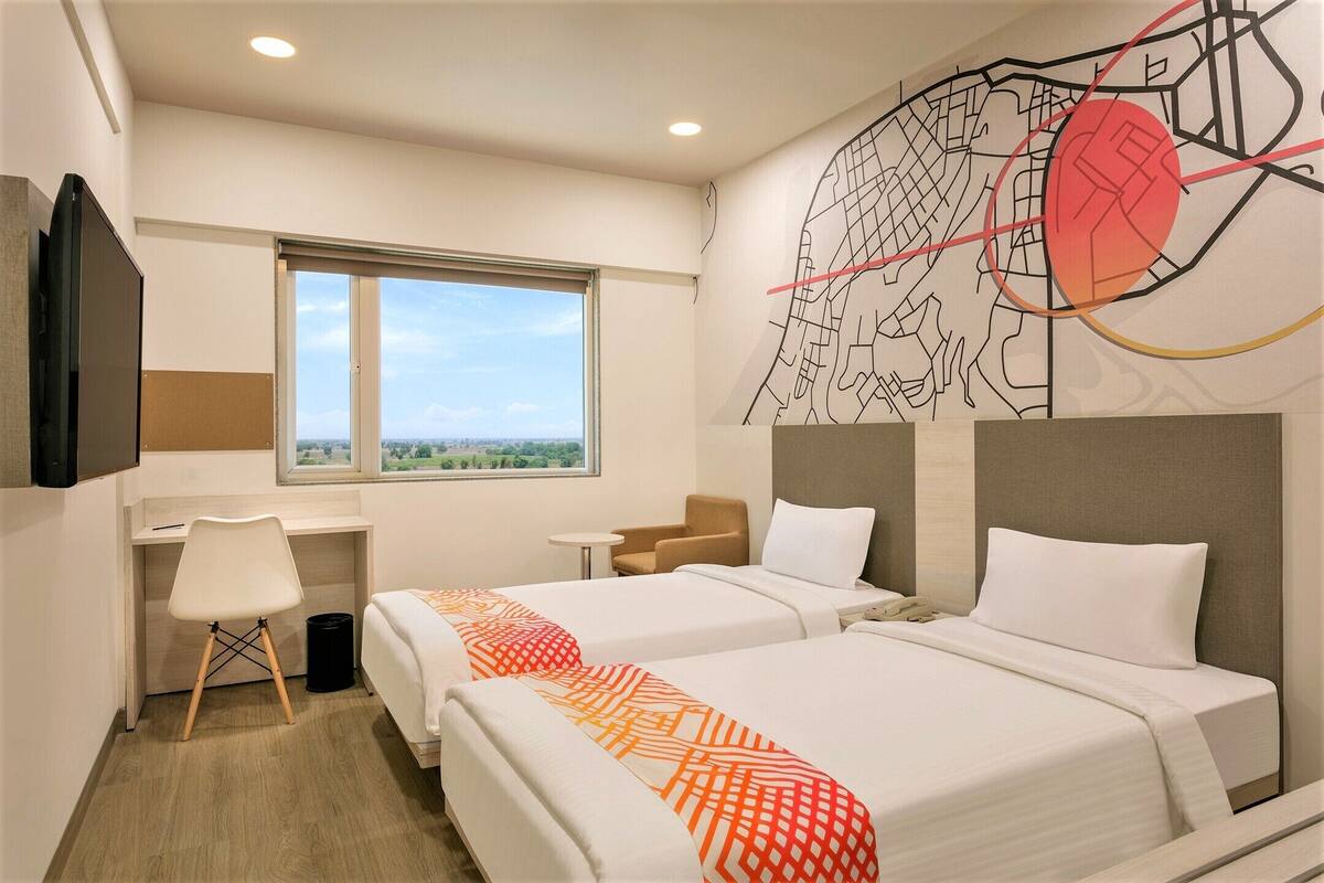 Quarto de hotel com duas camas, uma mesa, uma cadeira, uma televisão e uma janela com vista para a cidade.