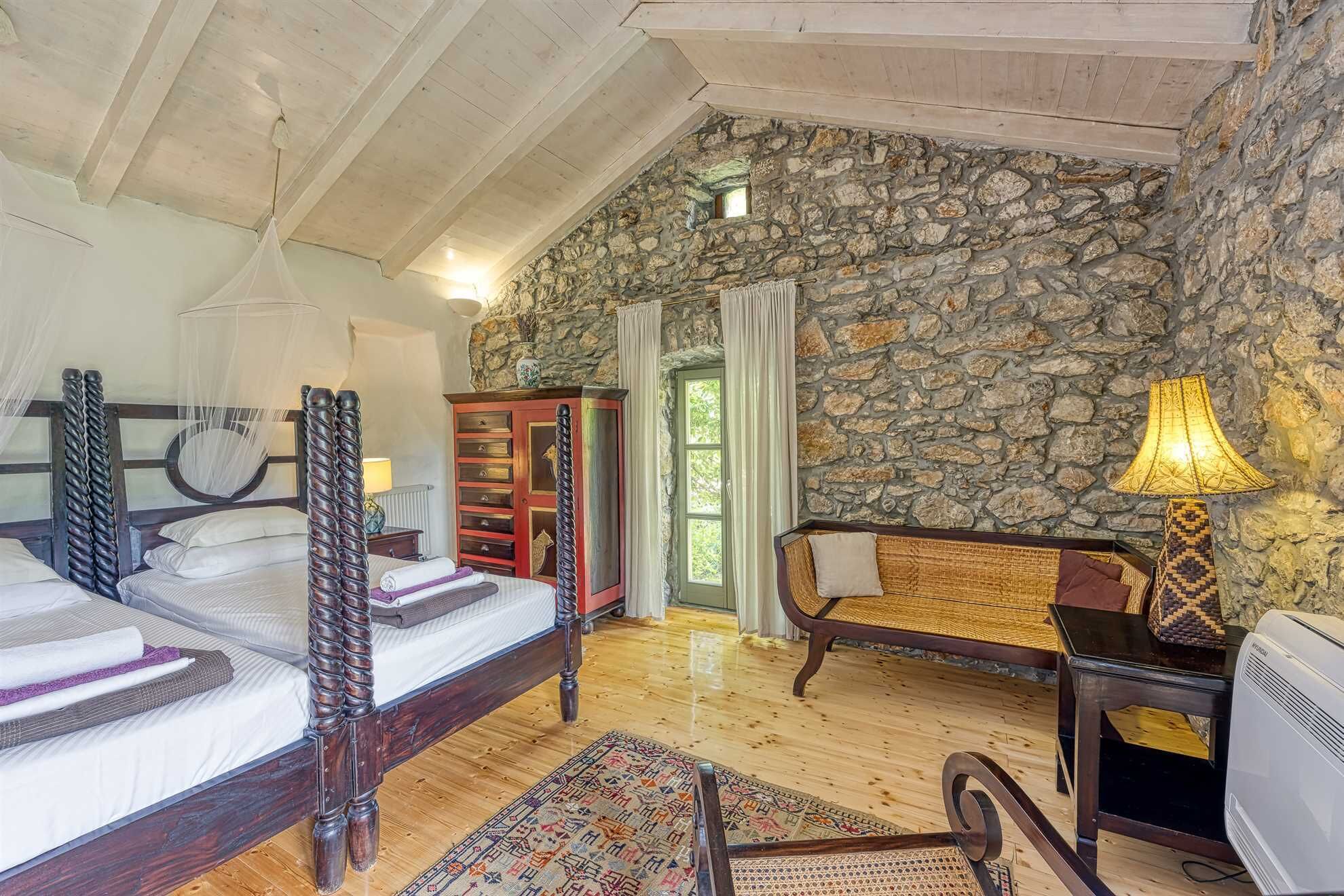 A bedroom with a stone wall, a bed with a canopy, a wooden chair, and a desk with a lamp.